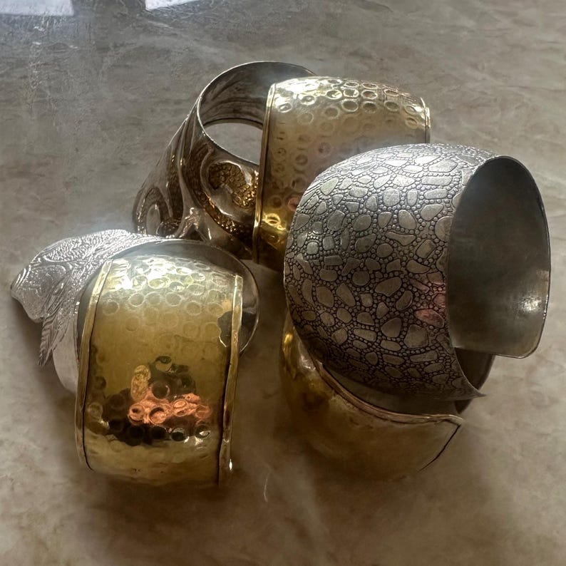 May include: An assortment of silver and gold-toned metal cuff bracelets. The bracelets feature various textures, including hammered and patterned designs. One bracelet has a detailed floral design, while others have a smooth, polished finish.