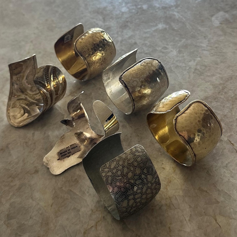 May include: An assortment of silver and gold-toned metal cuff bracelets. Some feature hammered textures, while others have intricate patterns. The bracelets are open-ended, allowing for easy wear.