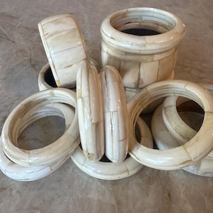 Może przedstawiać: Kolekcja kremowobiałych, segmentowych bransoletek i małego cylindrycznego pojemnika. Biżuteria ma naturalny, organiczny wygląd, z widocznymi liniami i zmiennymi kolorami. Bransoletki różnią się rozmiarem i kształtem, tworząc warstwowy efekt.