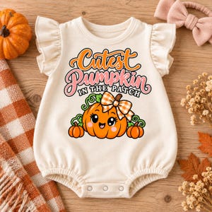 Pode incluir: Um macacão de bebé cor de creme com mangas com folhos, apresentando um gráfico de uma abóbora laranja sorridente com um laço xadrez e o texto Cutest Pumpkin In The Patch em letras cor de laranja e rosa, com duas abóboras pequenas.
