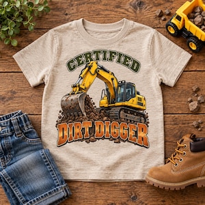 May include: A beige short-sleeve t-shirt featuring a graphic of a yellow excavator digging into a pile of dirt. The text above the excavator reads CERTIFIED in green, and the text below reads DIRT DIGGER in orange with a dark outline.