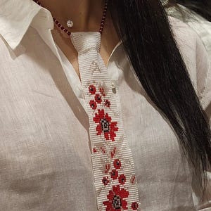 Könnte beinhalten: Eine weiße Perlen-Krawattenkette mit einem floralen Design in Rot und Schwarz. Die Halskette hat eine rote Perlenkette und einen kleinen, runden, funkelnden Ohrstecker. Die Krawatte zeigt ein detailliertes Muster aus Blumen und Blättern.