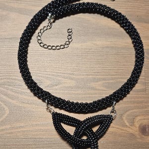 May include: A black beaded necklace featuring a circular rope-style chain and a central triangular triquetra pendant. The piece is constructed from small, uniform black beads and includes a silver-toned metal lobster clasp and an adjustable chain.