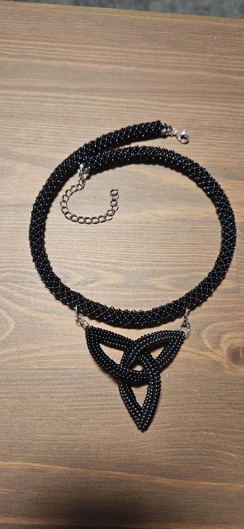May include: A black beaded necklace featuring a circular rope-style chain and a central triangular triquetra pendant. The piece is constructed from small, uniform black beads and includes a silver-toned metal lobster clasp and an adjustable chain.