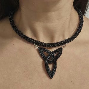 May include: A black beaded choker necklace featuring a central triangular pendant made of interconnected loops. The entire piece is constructed from small, uniform black beads, with silver-toned metal jump rings connecting the pendant to the choker.