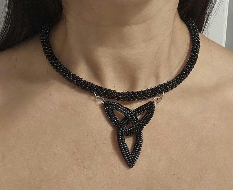 May include: A black beaded choker necklace featuring a central triangular pendant made of interconnected loops. The entire piece is constructed from small, uniform black beads, with silver-toned metal jump rings connecting the pendant to the choker.
