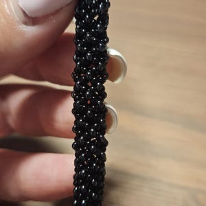 May include: A cylindrical bracelet crafted from small, glossy black beads woven into a dense, uniform tubular pattern. The surface of the accessory features a consistent, textured appearance created by the tightly linked, reflective dark glass beads.
