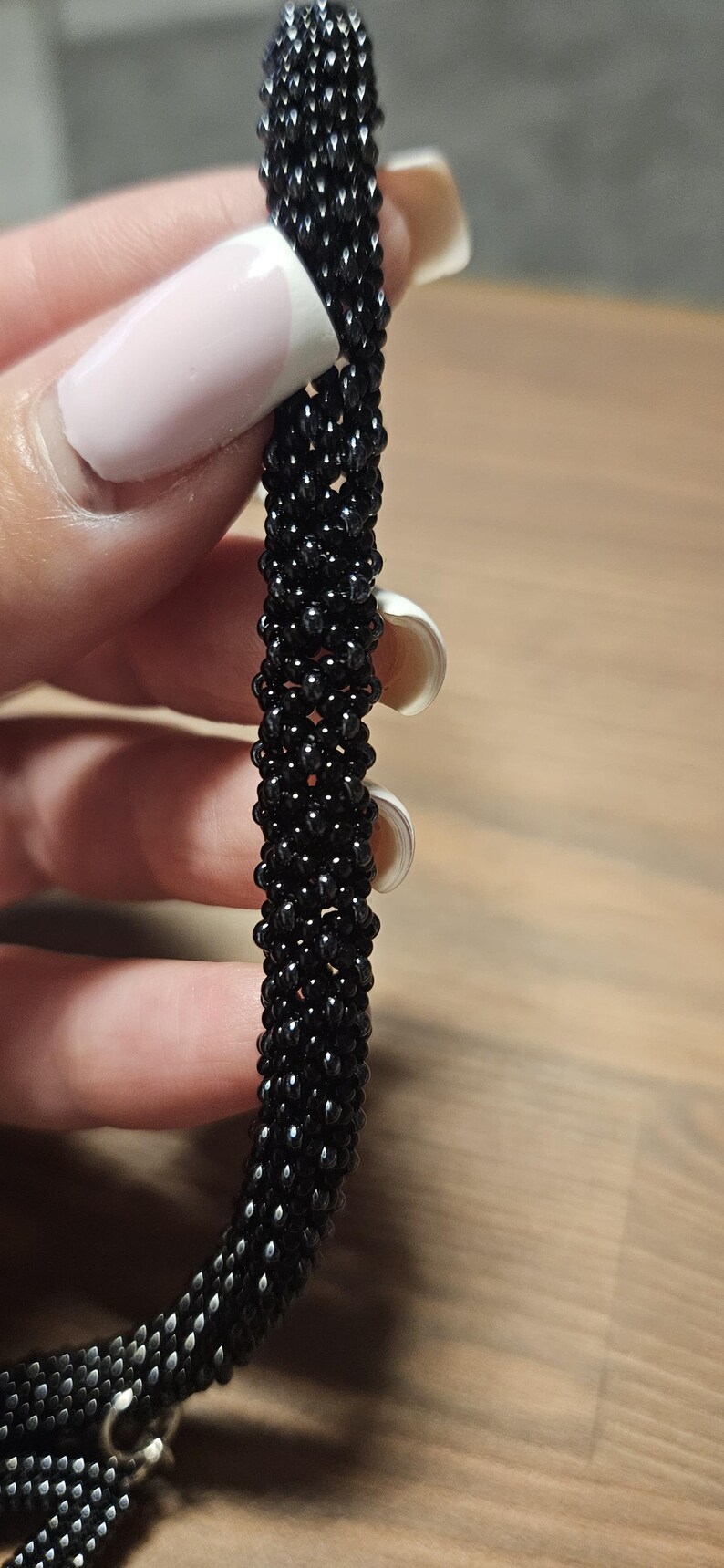 May include: A cylindrical bracelet crafted from small, glossy black beads woven into a dense, uniform tubular pattern. The surface of the accessory features a consistent, textured appearance created by the tightly linked, reflective dark glass beads.