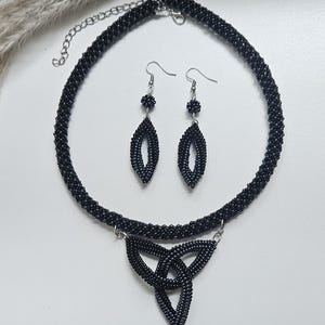 May include: A black beaded necklace and earring set. The necklace features a circular beaded band with a central triangular triquetra-style pendant. The matching earrings consist of a small beaded sphere above a larger, elongated, hollow diamond-shaped bead.