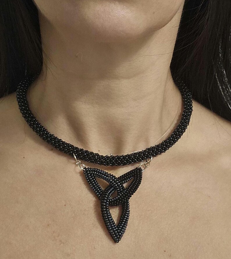 May include: A black beaded choker necklace featuring a central triangular pendant made of interconnected beaded loops. The necklace is constructed from small, uniform black beads and is attached to the pendant with silver-toned metal jump rings.