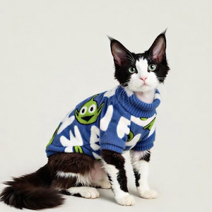 Könnte beinhalten: Eine schwarz-weiße Katze trägt einen blauen Pullover mit einem grünen Alien-Design. Die Katze sitzt und schaut in die Kamera. Der Pullover hat ein weißes Muster und einen gerippten Kragen.
