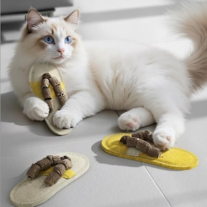 May include: A fluffy white cat with blue eyes lies next to three felt cat toys shaped like sandals. The sandals have a yellow sole and brown, stick-like accents. One sandal is held by the cat.