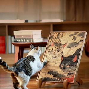 Könnte beinhalten: Ein Kratzbrett für Katzen aus Holz mit einer japanisch inspirierten Illustration von zwei Katzen mit orangefarbenen Augen und roten Akzenten. Eine Schildpattkatze interagiert mit dem Kratzbrett. Das Kratzbrett steht auf einem Holzständer, im Hintergrund Bücher auf einem kleinen Tisch.