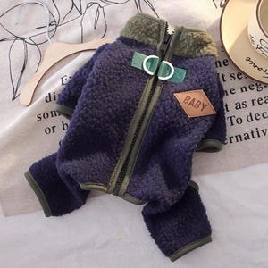 May include: A navy blue dog jacket with a green collar and trim. The jacket has a zipper closure and a small brown patch that says "BABY". The jacket is made of a soft, fuzzy material.