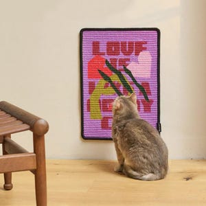 Könnte beinhalten: Ein rechteckiges Wandbild mit dem Wort "LOVE" in roten Buchstaben und abstrakten Blumendesigns in Rot, Grün und Gelb auf lila Hintergrund. Eine graue Katze sitzt auf dem Boden und blickt auf das Wandbild. Das Wandbild hat einen schwarzen Rand.