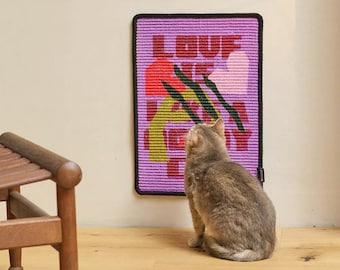 May include: A rectangular wall hanging with the word "LOVE" in red letters and abstract floral designs in red, green, and yellow on a purple background. A gray cat sits on the floor, looking up at the wall hanging. The wall hanging has a black border.