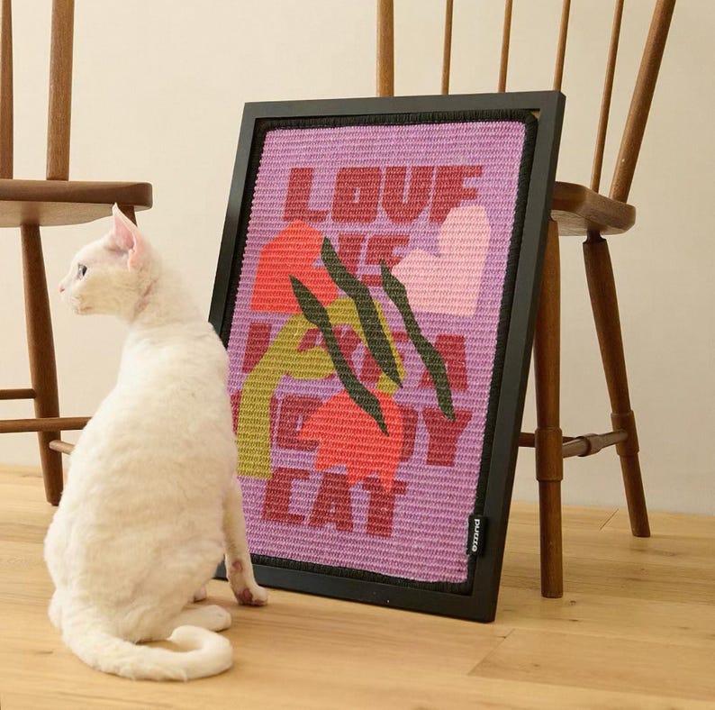 K&ouml;nnte beinhalten: Ein gerahmtes Kunstwerk mit dem Text "LOVE ME MY CAT" in roten Buchstaben auf rosa Hintergrund, mit bunten floralen Akzenten. Eine wei&szlig;e Katze sitzt auf dem Boden neben dem Kunstwerk. Der Rahmen ist schwarz.