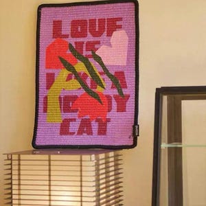 K&ouml;nnte beinhalten: Ein rechteckiger, violetter Wandbehang mit dem Text "LOVE ME LIKE A KITTY CAT" in roten Buchstaben. Das Design umfasst abstrakte Formen in Rot, Gelb und Rosa, mit schwarzen Kratzspuren. Die Kanten sind schwarz.