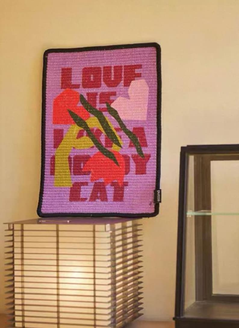 K&ouml;nnte beinhalten: Ein rechteckiger, violetter Wandbehang mit dem Text "LOVE ME LIKE A KITTY CAT" in roten Buchstaben. Das Design umfasst abstrakte Formen in Rot, Gelb und Rosa, mit schwarzen Kratzspuren. Die Kanten sind schwarz.