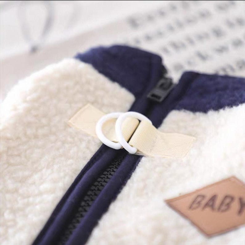 May include: Close-up of a cream-colored, fluffy fleece jacket with navy blue accents. The jacket features a black zipper, a beige strap with white rings, and a brown patch that reads "BABY".