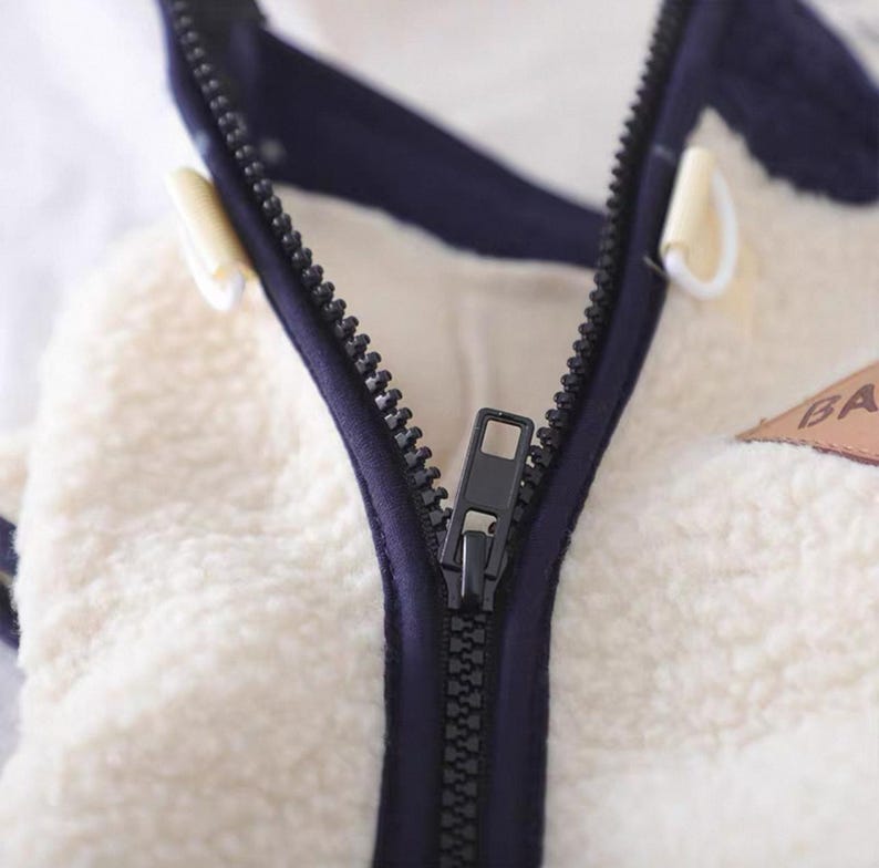 May include: Close-up of a cream-colored, fluffy jacket with a navy blue trim and a black zipper. The jacket has toggle closures and a small patch with the letters "BA".