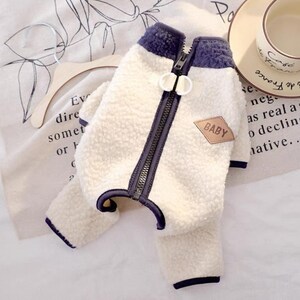 May include: A cream-colored dog jacket with a navy blue trim and zipper. The jacket has a soft, fuzzy texture and a small patch that says "BABY". The jacket is designed to keep a dog warm and comfortable.