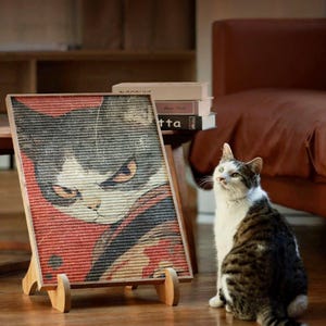 Könnte beinhalten: Ein gerahmtes Katzenporträt auf einer Holzstaffelei. Das Kunstwerk zeigt eine Nahaufnahme des Gesichts einer Katze in Grau-, Weiß- und Rottönen. Eine echte Katze mit ähnlichen Markierungen sitzt in der Nähe und blickt nach oben.