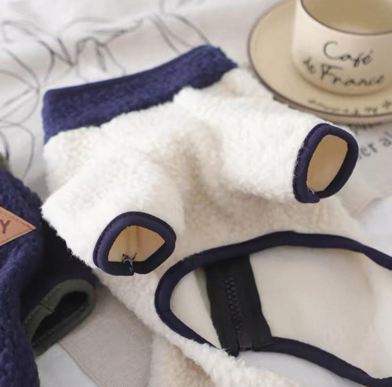 May include: A white and navy blue dog sweater with a zipper closure. The sweater has navy blue trim around the collar and sleeves. The material appears to be soft and fluffy.