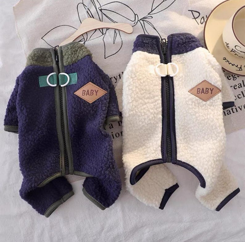 May include: Two dog jackets, one navy blue and one cream-colored, are displayed. Both jackets have a fuzzy texture, a zipper closure, and a small patch that reads "BABY". The jackets are designed to keep small dogs warm.