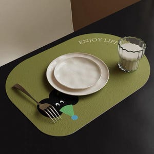 Könnte beinhalten: Eine ovale grüne Tischmatte mit einem Cartoon-Charakter-Design, zwei weißen Tellern, einem Glas Milch und einer Gabel. Auf der Tischmatte steht "ENJOY LIFE". Die Cartoonfigur hat einen schwarzen Kopf, weiße Augen und einen grünen und blauen Hut.