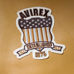 Puede incluir: Parche bordado con la palabra "AVIREX" en la parte superior, un dise&ntilde;o de escudo con estrellas y rayas, y las palabras "ESTABLISHED NY 1975". El parche es blanco con detalles en marr&oacute;n, dorado y blanco, sobre un fondo beige.