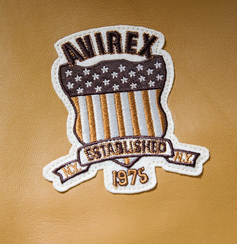 Puede incluir: Parche bordado con la palabra "AVIREX" en la parte superior, un dise&ntilde;o de escudo con estrellas y rayas, y las palabras "ESTABLISHED NY 1975". El parche es blanco con detalles en marr&oacute;n, dorado y blanco, sobre un fondo beige.