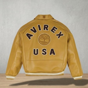 Puede incluir: Chaqueta de cuero amarillo mostaza con la palabra "AVIREX" arqueada en la espalda, encima de "USA". Un emblema circular con un dise&ntilde;o de &aacute;rbol est&aacute; centrado entre las palabras. La chaqueta tiene cuello y pu&ntilde;os y dobladillo a rayas blancas y crema.