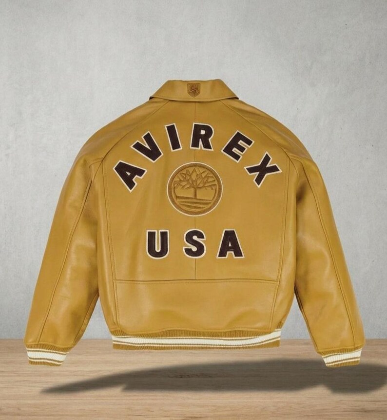 Puede incluir: Chaqueta de cuero amarillo mostaza con la palabra "AVIREX" arqueada en la espalda, encima de "USA". Un emblema circular con un dise&ntilde;o de &aacute;rbol est&aacute; centrado entre las palabras. La chaqueta tiene cuello y pu&ntilde;os y dobladillo a rayas blancas y crema.