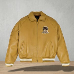 Puede incluir: Chaqueta de cuero color mostaza con cuello y pu&ntilde;os acanalados. La chaqueta tiene cierre de cremallera y un parche en el pecho con el texto "AVIREX 1975". Los pu&ntilde;os y el dobladillo tienen rayas blancas.
