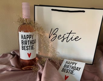 May include: A bottle of rosé wine with a label that reads "HAPPY BIRTHDAY BESTIE" and a red heart. A gift bag with the word "Bestie" and a card with the same message are also present. Gold tinsel surrounds the bottle.