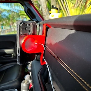 Puede incluir: Un soporte rojo para tel&eacute;fono instalado en un veh&iacute;culo, que sostiene un tel&eacute;fono inteligente negro. Un patito de goma amarillo con un sombrero negro est&aacute; sentado en el salpicadero. El interior presenta cuero negro y un cable de carga blanco.