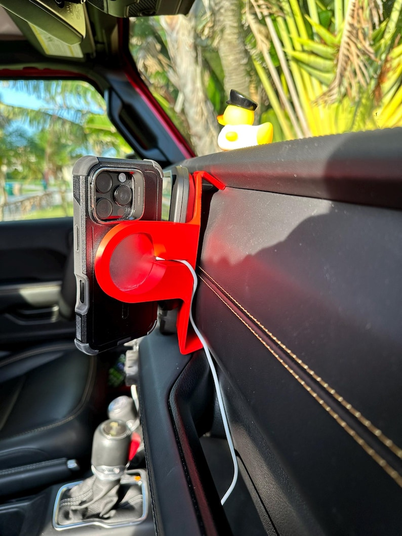 Puede incluir: Un soporte rojo para tel&eacute;fono instalado en un veh&iacute;culo, que sostiene un tel&eacute;fono inteligente negro. Un patito de goma amarillo con un sombrero negro est&aacute; sentado en el salpicadero. El interior presenta cuero negro y un cable de carga blanco.