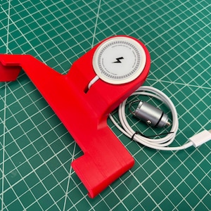 Puede incluir: Un soporte para tel&eacute;fono rojo con un cargador inal&aacute;mbrico blanco y un cable de carga blanco. El cargador tiene un s&iacute;mbolo de rayo negro. El soporte est&aacute; sobre una superficie de cuadr&iacute;cula verde.