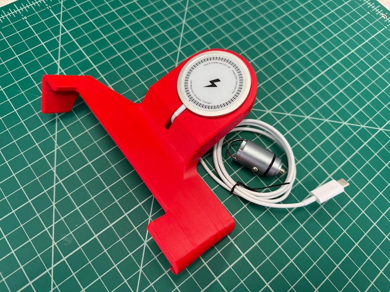 Puede incluir: Un soporte para tel&eacute;fono rojo con un cargador inal&aacute;mbrico blanco y un cable de carga blanco. El cargador tiene un s&iacute;mbolo de rayo negro. El soporte est&aacute; sobre una superficie de cuadr&iacute;cula verde.
