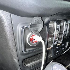 Puede incluir: Primer plano del interior de un coche con un salpicadero negro, un puerto de carga USB y un cable de carga blanco enchufado. El puerto tiene un interior rojo y una cubierta protectora. Se ven otros controles.