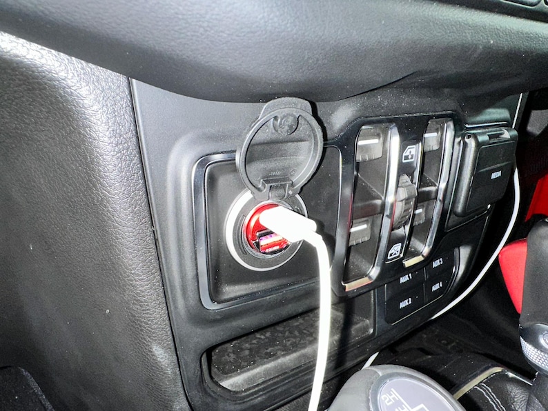 Puede incluir: Primer plano del interior de un coche con un salpicadero negro, un puerto de carga USB y un cable de carga blanco enchufado. El puerto tiene un interior rojo y una cubierta protectora. Se ven otros controles.