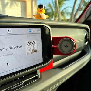 Puede incluir: Interior del salpicadero de un coche con una pantalla digital que muestra los controles de m&uacute;sica y la car&aacute;tula del &aacute;lbum. Un peque&ntilde;o pato de goma con sombrero de copa est&aacute; sentado en el salpicadero. Un cargador de tel&eacute;fono rojo est&aacute; montado en el salpicadero.
