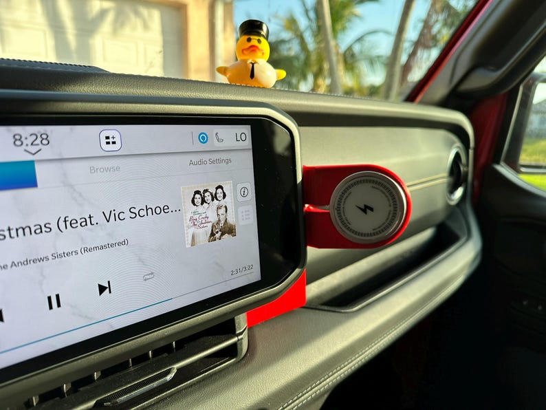 Puede incluir: Interior del salpicadero de un coche con una pantalla digital que muestra los controles de m&uacute;sica y la car&aacute;tula del &aacute;lbum. Un peque&ntilde;o pato de goma con sombrero de copa est&aacute; sentado en el salpicadero. Un cargador de tel&eacute;fono rojo est&aacute; montado en el salpicadero.