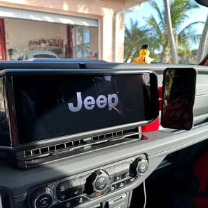 Puede incluir: Vista interior del salpicadero de un veh&iacute;culo Jeep. La gran pantalla de infoentretenimiento muestra el logotipo de Jeep. Un smartphone est&aacute; montado a la derecha. Un patito de goma con sombrero est&aacute; en el salpicadero. El salpicadero es negro y rojo.