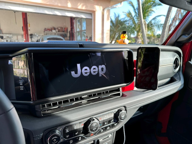 Puede incluir: Vista interior del salpicadero de un veh&iacute;culo Jeep. La gran pantalla de infoentretenimiento muestra el logotipo de Jeep. Un smartphone est&aacute; montado a la derecha. Un patito de goma con sombrero est&aacute; en el salpicadero. El salpicadero es negro y rojo.
