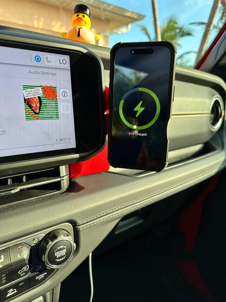 Puede incluir: Un tel&eacute;fono inteligente en una funda negra se est&aacute; cargando en un coche. El tel&eacute;fono muestra un s&iacute;mbolo de carga verde y 81% cargado. El salpicadero del coche incluye ajustes de audio y un patito de goma.