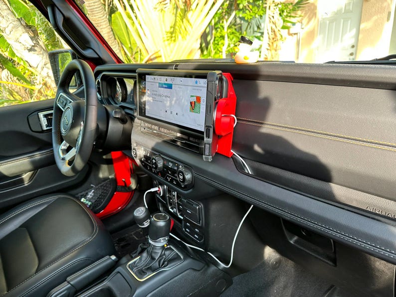Puede incluir: Vista interior de un Jeep Wrangler rojo. El salpicadero cuenta con una gran pantalla de infoentretenimiento, un soporte rojo para el tel&eacute;fono y varios controles. Un patito de goma est&aacute; en el salpicadero. El volante y los asientos son negros.