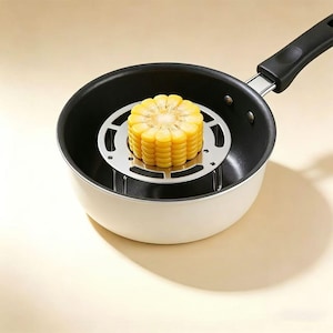 May include: A stainless steel circular steaming rack with three legs sits inside a white saucepan with a black interior and a black handle. A single yellow corn cob segment is placed upright on the center of the metal rack for cooking.
