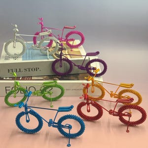 May include: A collection of colorful, miniature wire bicycles. The bikes are in various colors, including silver, pink, purple, green, blue, red, and yellow. Each bike has a unique design and is made of coiled wire. The bikes are displayed on a white surface.